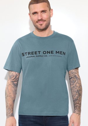 Mężczyzna z krótkimi włosami i tatuażami na całym ramieniu nosi niebieską koszulkę z napisem "STREET ONE MEN ORIGINAL SUPPLY CO." na jednolitym tle.