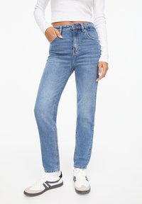 Högmidjade blå denimjeans med rak passform, utrustade med fem fickor, bälteshällor och fransade fållkanter. Matchas med vita sneakers.