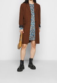 Hnědý kabát ze směsi vlny s klopami a dvěma předními kapsami, vrstvený přes modré květinové šaty, doplněný černými robustními kotníkovými botami a béžovou kabelkou.