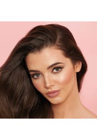 Junge Frau mit langen braunen Haaren, klarer Haut, markanten Augenbrauen, Katzenaugeneyeliner und mattem pinkfarbenem Lippenstift vor einem sanften rosa Hintergrund.