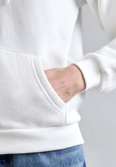 Sweatshirt blanc avec une poche kangourou, des poignets et un ourlet côtelés. Le tissu semble doux et texturé, ajusté au poignet, en contraste avec un jean bleu.