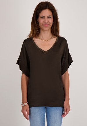Bruine V-halsblouse met korte mouwen, zachte stof en decoratieve bies langs de halslijn. Gecombineerd met blauwe spijkerbroek.