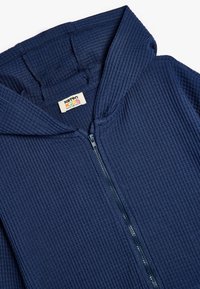 Mørkeblå hættetrøje med lynlås lavet af ribbet stof, med hætte, der har et mærke med teksten "Karton Kids" og en robust lynlås.