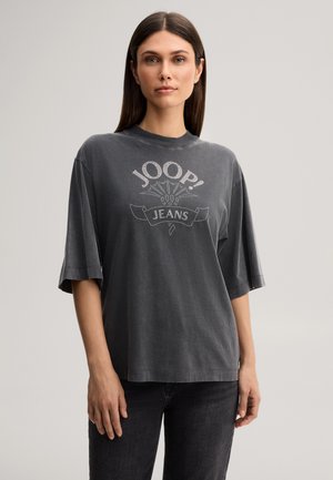 Młoda kobieta z prostymi brązowymi włosami, ubrana w luźny ciemnoszary t-shirt z napisem "JOOP! JEANS" i czarne jeansy, stojąca na neutralnym tle.