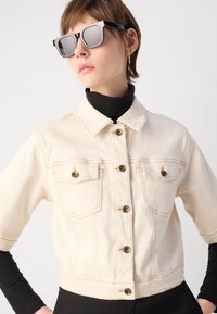 Krämfärgad kort denimjacka med knappstängning, två bröstfickor och synliga sömnadsdetaljer. Bärs över en svart polotröja.