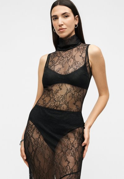 Femme aux longs cheveux foncés portant une robe noire en dentelle transparente sans manches et des sous-vêtements noirs, posant avec une main sur la hanche.