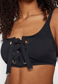 Svart bikini topp med snøring foran og store øyer, laget av et glatt materiale. Utsmykket med metallkomponenter for ekstra detaljer.