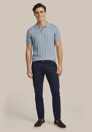 Lachende man staand, draagt een lichtblauw poloshirt met korte mouwen, donkerblauwe broek, bruine riem en donkerbruine instapschoenen op een beige achtergrond.