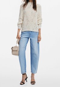 Cremefarbener Strickpullover mit bunten Sprenkeln, lockerer Passform und gerippten Bündchen. Kombiniert mit hellblauen High-Waist-Jeans und schwarzen Sandalen mit Absatz.