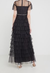 Mujer vestida con un largo vestido de malla negra con volantes y un detalle en la cintura de flores, combinado con sandalias de tacón alto plateadas, de espaldas.