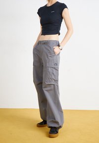 Černé zkrácené tričko s nařasenými stranami, šedé cargo kalhoty s bočními kapsami a logem, a černé tenisky s gumovou podrážkou, stojící na žluté podlaze.