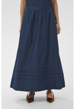 Žena oblečená v dlhej tmavomodrej rifľovej sukni s skladanými detailmi pri okraji a viditeľnými hnedými špicatými podpätkami pod ňou.