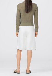 Kvinna med långt rakt hår iklädd en olivgrön stickad tröja, vita knälånga shorts och beigea slip-on sandaler, vänd bort.