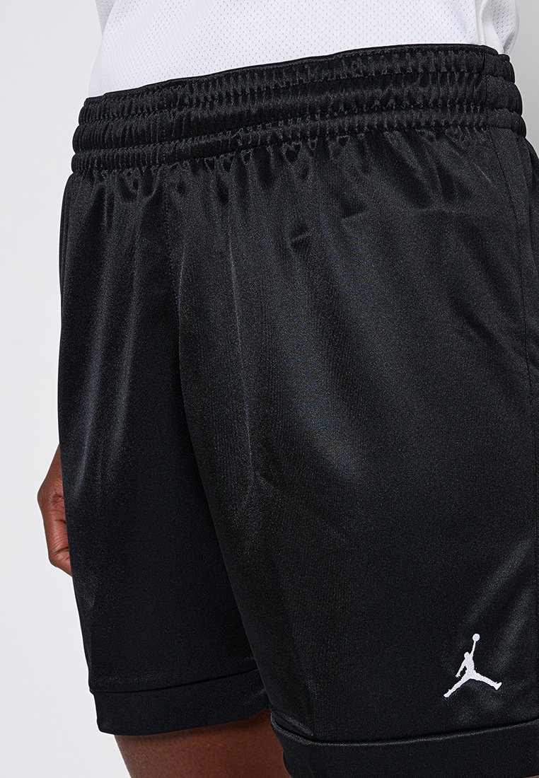 Shorts de sport noirs avec taille élastique et logo Jumpman blanc portés par une personne à la peau marron et portant un haut blanc.