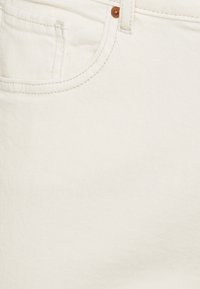 Valkeat farkut, joissa on teksturoitu pinta, pienellä etutaskulla ja erottuvalla ruskealla napilla. Saumoissa näkyy ompelu.