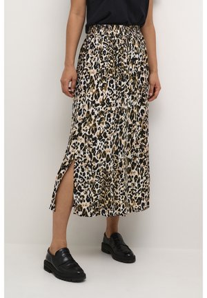 Femme portant une jupe midi imprimé léopard avec une fente latérale, un haut noir et des mocassins noirs, debout devant un fond uni.
