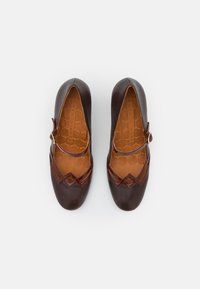 Chaussures en cuir marron avec un bout arrondi, présentant un design découpé décoratif et une sangle avec une boucle dorée. Semelle intérieure texturée visible.