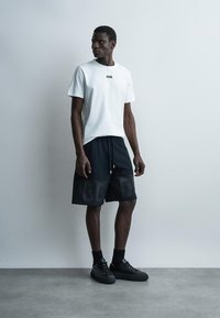 Homme debout contre un mur gris clair portant un t-shirt blanc GCDS, un short noir avec le logo GCDS, des chaussettes noires et des baskets noires.