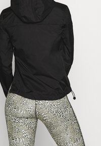 Sort hættejakke med indsnøret talje, kombineret med mønstrede grønne og hvide leggings med leopardprint.