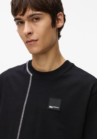 Sweatshirt negru cu guler rotund, având cusături albe contrastante și un patch logo pătrat pe piept. Textură moale, design casual.