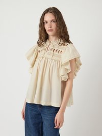 Cream blouse with flutter sleeves, textured embroidered yoke, and gathered waistline. Paired with blue jeans for a relaxed fit.