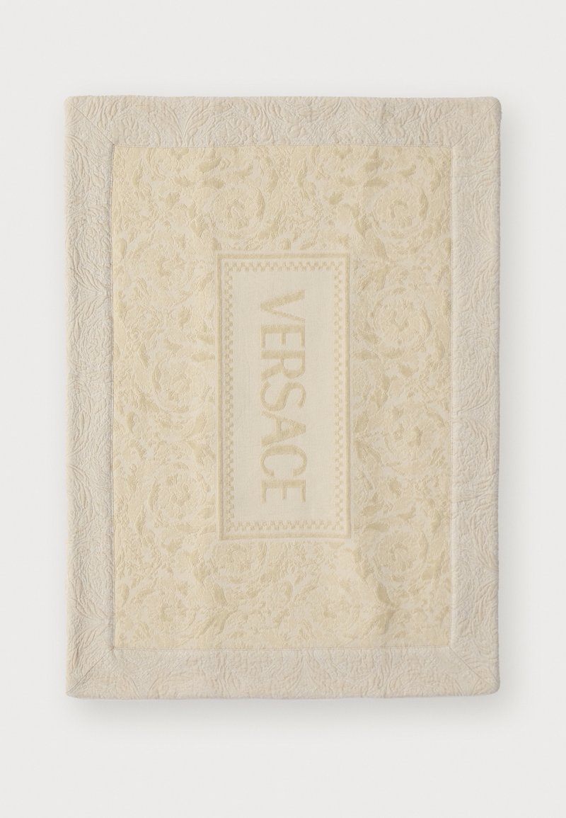 Versace blanket in soft cream with an embossed floral pattern, featuring a rectangular logo patch in lighter cream centered on the fabric.