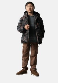 Dziecko w wzorzystej czarnej zimowej kurtce, szarej swetrze, brązowych spodniach cargo, brązowych sneakersach i dzianinowej czapce, spoglądające lekko w górę i w prawo.