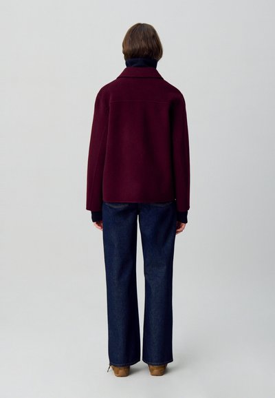 Chaqueta de mezcla de lana color burdeos con cuello ancho, corte holgado y puños en azul marino, combinada con vaqueros acampanados de color azul oscuro y calzado marrón.