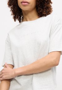 Γυναίκα με σγουρά μαλλιά φοράει ανοιχτό γκρι κοντομάνικο πουκάμισο με διακριτικό κείμενο "Tommy Hilfiger", κρατώντας το ένα χέρι της σταυρωτά πάνω από το σώμα της.