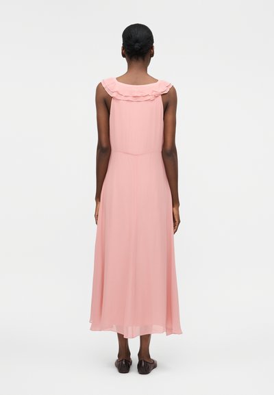 Γυναίκα με σκούρο δέρμα και μαλλιά δεμένα σε κότσο, που φοράει ένα αμάνικο, ανοιχτό ροζ φόρεμα μέχρι τους αστραγάλους με σφηκοφωλιά στη λαιμόκοψη, στέκεται γυρισμένη από πίσω.