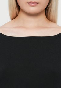 Close-up van de nek en schouders van een vrouw die een zwarte off-shoulder top draagt, met steil blond haar zichtbaar.