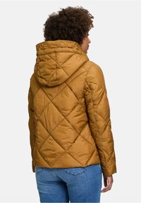 Vatteret, sennepsfarvet jakke med hætte, der har diagonale søm mønstre og en tætsiddende silhuet. Båret over blå jeans.