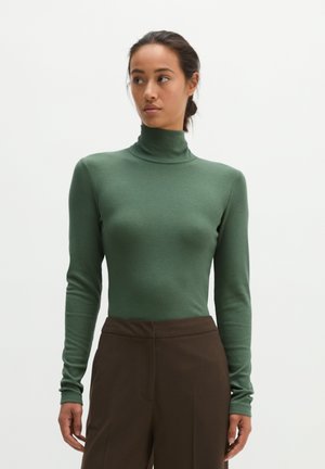 Femme portant un pull vert moulant à col roulé et un pantalon marron taille haute, debout devant un fond blanc uni.