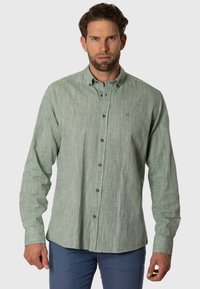 Camisa de botones verde claro con una tela texturizada, que presenta un pequeño logo bordado y botones marrones, llevada sobre pantalones azules.