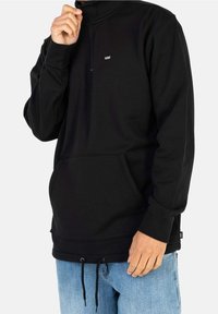 Uomo che indossa una felpa nera a mezza zip Vans con tasca frontale e cordini, abbinata a jeans blu chiaro, su uno sfondo semplice.