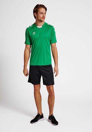 Groene sportshirt met korte mouwen, zwarte shorts en zwarte sneakers. Het shirt heeft witte accenten en een logo op de borst.