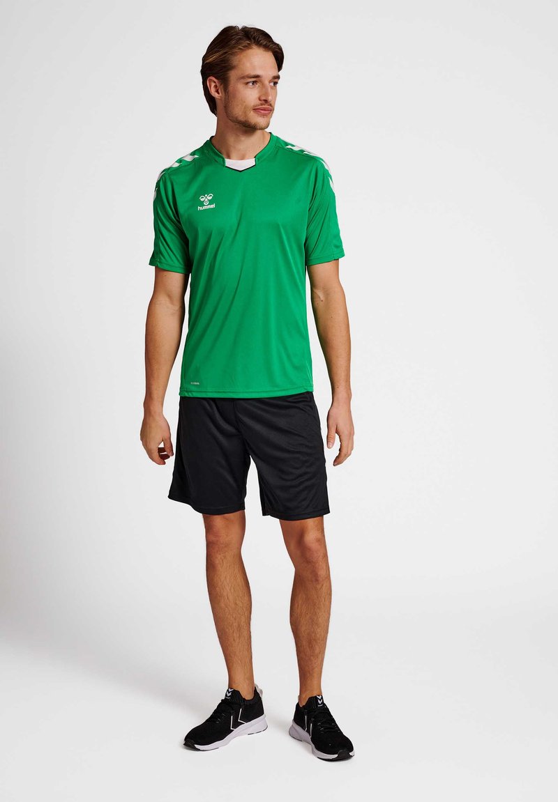 Grünes Sportshirt mit kurzen Ärmeln, schwarze Shorts und schwarze Sneaker. Das Shirt hat weiße Akzente und ein Logo auf der Brust.