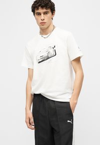 Νέος με σγουρά μαλλιά, φορώντας λευκό μπλουζάκι με γραφικό αυτοκινήτου και μαύρο παντελόνι Puma, ποζάρει με το ένα χέρι στην τσέπη.