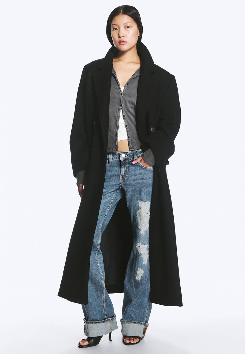 Femme portant un long manteau noir, une chemise grise boutonnée par-dessus un crop top blanc, un jean bleu usé avec des revers roulés, et des talons noirs ouverts.