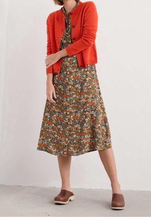 Kvinde iført en orange cardigan med knapper over en blomsterprintet midikjole med brune lædertræsko, stående foran en ensfarvet baggrund.