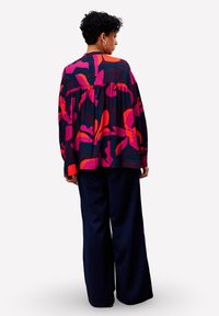 Personne avec des cheveux bouclés courts portant un haut ample à motifs floraux colorés et un pantalon large sombre, de dos sur un fond blanc.