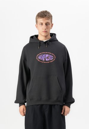 Joven con una sudadera negra con capucha y un logotipo colorido de Rip Curl acompañado del texto "Raw Energy", de pie contra un fondo blanco liso.