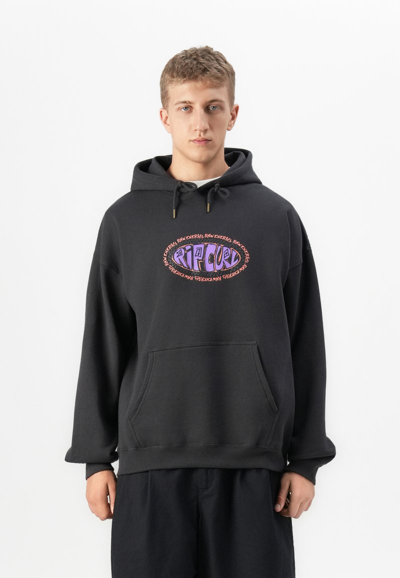 Jonge man in een zwarte hoodie met een kleurrijk Rip Curl-logo en de tekst "Raw Energy" die tegen een effen witte achtergrond staat.