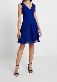 Niebieska koronkowa sukienka z dekoltem w kształcie litery V, dopasowaną talią i rozkloszowanym dołem. Posiada kwiatowe wzory i przezroczyste detale. Noszona z czarnymi, odkrytymi szpilkami.