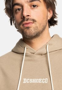 Beige hoodie met een trekkoord en witte borduursel "DCSHOECO" op de borst. Het materiaal lijkt zacht en gestructureerd.