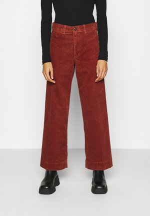 Person wearing rust-red wide-leg corduroy pants, black long-sleeve top, and black chunky ankle boots against a plain background.