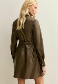 Blonde vrouw in een bruin leren jurk met ceintuur en lange mouwen, staand zijwaarts tegen een lichtbeige muur.