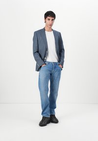 Grå mönstrad blazer över en vit t-shirt, tillsammans med ljusblå jeans och svarta sneakers. Modellen står med händerna i fickorna, framifrån.