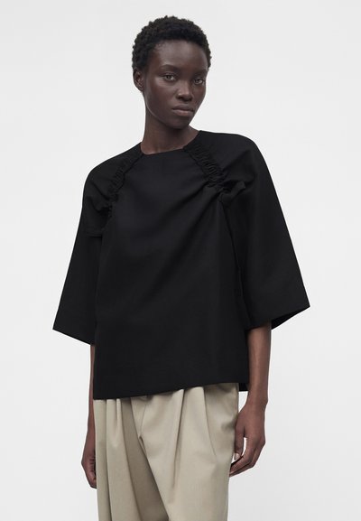 Άτομο με σκουρόχρωμο δέρμα που φοράει ένα φαρδύ μαύρο τοπ με ραμμένη τσάκιση και φαρδιά μανίκια, σε συνδυασμό με μπεζ πλισέ ψηλόμεσα παντελόνια.