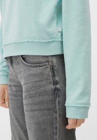 Primer plano de una persona que lleva un sudadera azul claro con puños y dobladillo acanalados, combinada con unos vaqueros desgastados de color negro.